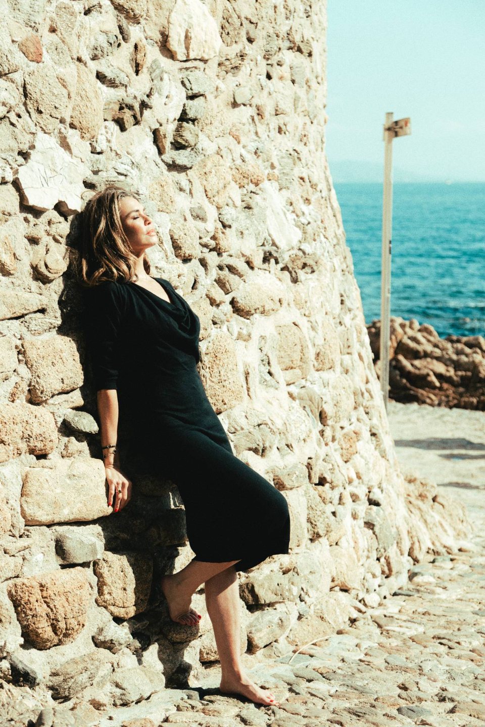 Tara leaning against a stone tower by the sea, eyes closed, her face turned to the sun.