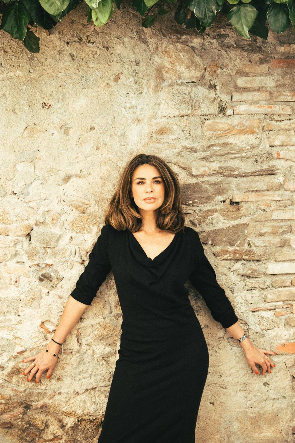 Tara in a black dress, leaning back against an old stone wall beneath ivy, looking at the camera.